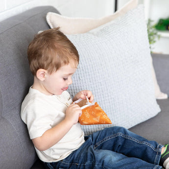 A young boy sits on a gray couch, peeking inside an orange Bumkins Reusable Snack Bag, Small 2-Pack: Sunshine and Grounded.