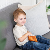 A toddler on a gray sofa smiles, holding an orange Bumkins Reusable Snack Bag, Small 2-Pack: Sunshine and Grounded.