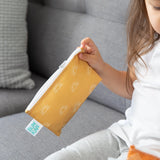A child on a grey couch holds a yellow Bumkins Reusable Snack Bag, Small 2-Pack: Sunshine and Grounded.