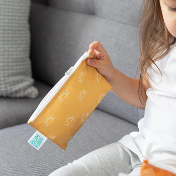 A child on a grey couch holds a yellow Bumkins Reusable Snack Bag, Small 2-Pack: Sunshine and Grounded.
