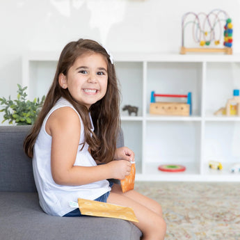 A smiling girl sits on a sofa with Bumkins Reusable Snack Bag, Small 2-Pack: Sunshine and Grounded. Shelves with toys are in the background.