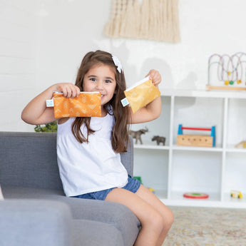 A girl smiles on a gray couch, holding Bumkins Reusable Snack Bag, Small 2-Pack: Sunshine and Grounded. A play area with toys is behind her.