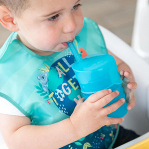 A toddler uses a Bumkins Silicone Straw Cup with Lid: Blue, drinking independently indoors while wearing a bib.