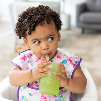 A toddler sips from a Bumkins Silicone Straw Cup with Lid: Sage, seated comfortably in a high chair.