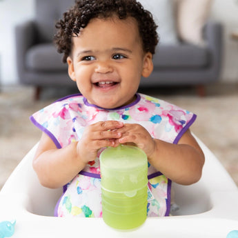 A smiling toddler sits in a high chair, wearing a colorful bib and holding a Bumkins Silicone Straw Cup with Lid: Sage.