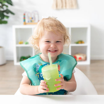 A toddler joyfully holds a Bumkins Silicone Straw Cup with Lid: Sage at a white table.