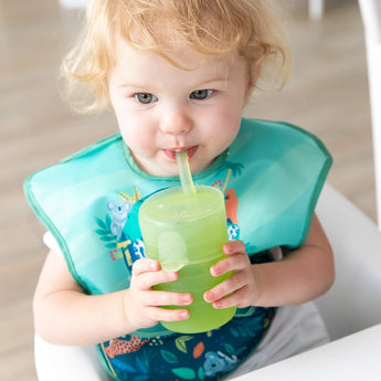 A toddler with blonde hair sips from a Bumkins Silicone Straw Cup with Lid: Sage, wearing a colorful bib with animal designs.