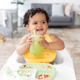 A baby in a high chair, wearing a yellow bib, holds a Bumkins Silicone Straw Cup with Lid: Sage, with food on a divided plate in front.