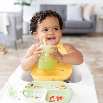A baby in a high chair, wearing a yellow bib, holds a Bumkins Silicone Straw Cup with Lid: Sage, with food on a divided plate in front.