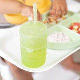 Tiny hands reach for food on a green divided plate as a Bumkins Silicone Straw Cup with Lid: Sage sits nearby.