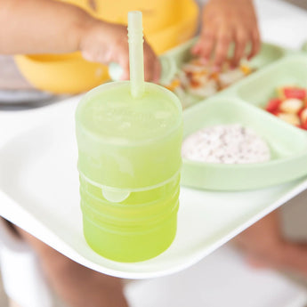 Tiny hands reach for food on a green divided plate as a Bumkins Silicone Straw Cup with Lid: Sage sits nearby.