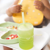 A toddler sits in a high chair with a Bumkins Silicone Straw Cup with Lid: Sage and a divided plate filled with treats.