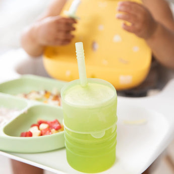 A toddler sits in a high chair with a Bumkins Silicone Straw Cup with Lid: Sage and a divided plate filled with treats.