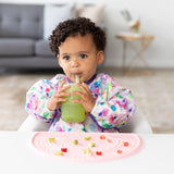 A toddler with a colorful bib sips from a Bumkins Silicone Straw Cup with Lid: Sage beside a pink mat with chopped fruits and veggies.