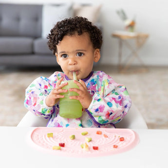 A toddler with a colorful bib sips from a Bumkins Silicone Straw Cup with Lid: Sage beside a pink mat with chopped fruits and veggies.