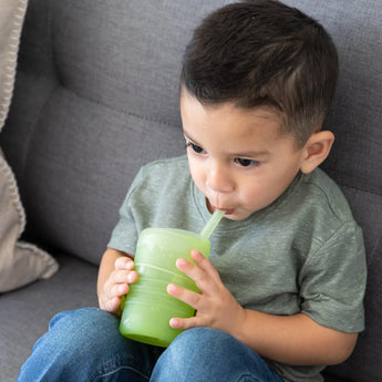 A child in a gray shirt sits on a couch, sipping from a Bumkins Silicone Straw Cup with Lid: Sage.