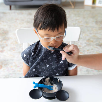 A child happily enjoys yogurt, being fed by an adult with a Bumkins Silicone Dipping Spoons: Mickey Mouse (Black and Blue) from a bowl.