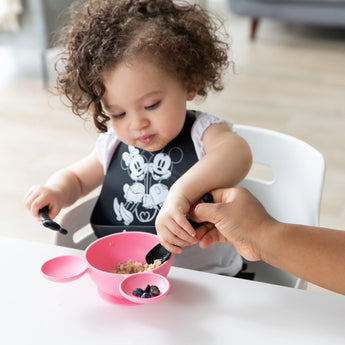 A toddler enjoys a meal from a pink bowl with Bumkins Silicone Dipping Spoons: Mickey Mouse (Classic Black), assisted by an adult's hand.