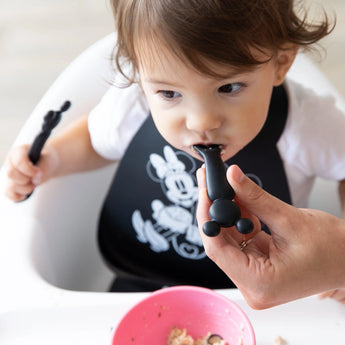 A toddler in a high chair with a bib is fed by an adult hand with a Bumkins Silicone Dipping Spoons: Mickey Mouse (Classic Black).