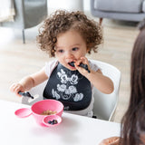 A curly-haired toddler in a bib enjoys blueberries from a pink bowl, using Bumkins Silicone Dipping Spoons: Mickey Mouse (Classic Black).
