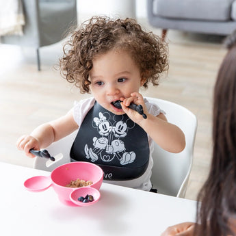 A curly-haired toddler in a bib enjoys blueberries from a pink bowl, using Bumkins Silicone Dipping Spoons: Mickey Mouse (Classic Black).