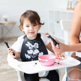 A toddler in a high chair in a bib holds a Bumkins Silicone Dipping Spoons: Mickey Mouse (Classic Black), while an adult assists in feeding.