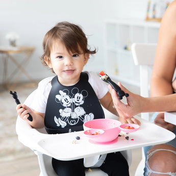 A toddler in a high chair in a bib holds a Bumkins Silicone Dipping Spoons: Mickey Mouse (Classic Black), while an adult assists in feeding.