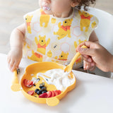 A child enjoys yogurt with fruit on a bear-shaped plate, using Bumkins Silicone Dipping Spoons: Winnie the Pooh with adult assistance.
