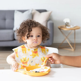 A curly-haired toddler eats with adult help from a dish with Bumkins Silicone Dipping Spoons: Winnie the Pooh, food smudged on their cheeks.
