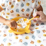 A child eats yogurt with Bumkins Silicone Dipping Spoons: Winnie the Pooh from a bear-shaped plate on a tablecloth, assisted by an adult.