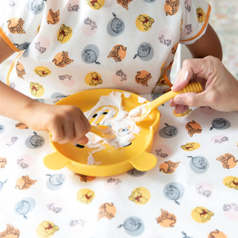 A child eats yogurt with Bumkins Silicone Dipping Spoons: Winnie the Pooh from a bear-shaped plate on a tablecloth, assisted by an adult.