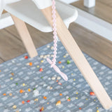 A pink pacifier clip hangs above a patterned mat scattered with Bumkins Silicone Dipping Spoons: Lollipop and food pieces.