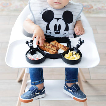 A baby in a high chair enjoys toast, eggs, and fruit using Bumkins Silicone Chewtensils®: Mickey Mouse and a Mickey-themed plate.