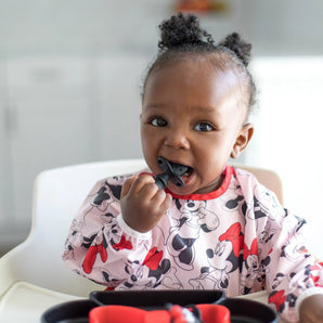A toddler in a Minnie Mouse bib is happily chewing on a Bumkins Silicone Chewtensils®: Minnie Mouse in a high chair.
