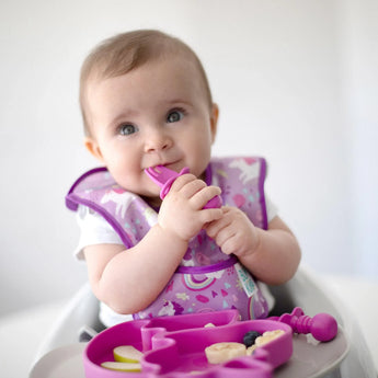 A baby in a high chair, ready for baby-led weaning, uses Bumkins Silicone Chewtensils® in fuchsia with a matching bib and spoon.