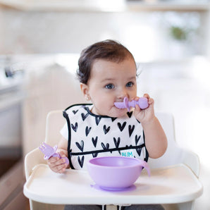 A toddler wearing a heart-patterned bib in a high chair eats from a purple bowl using Bumkins Silicone Chewtensils®: Lavender.