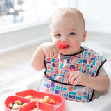 Baby enjoys baby-led weaning with a colorful bib and a red divided plate, exploring textures using Bumkins Silicone Chewtensils®.