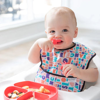 Baby enjoys baby-led weaning with a colorful bib and a red divided plate, exploring textures using Bumkins Silicone Chewtensils®.