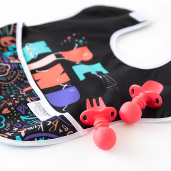 On a white surface lies a colorful baby bib with abstract designs paired with Bumkins Silicone Chewtensils®: Red.