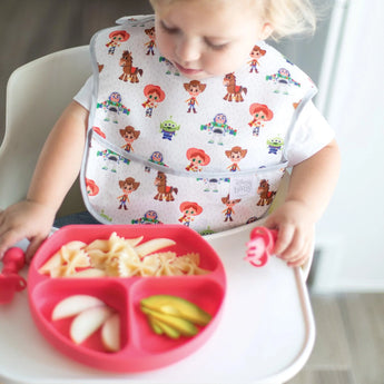 A toddler in a highchair using Silicone Chewtensils®: Red by Bumkins and eyeing a divided plate of food while wearing a cartoon bib.