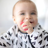 A smiling baby wears a heart-patterned bib, holding Bumkins Silicone Chewtensils®: Red with sensory bumps.