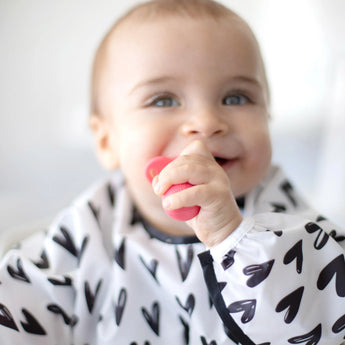 A smiling baby wears a heart-patterned bib, holding Bumkins Silicone Chewtensils®: Red with sensory bumps.