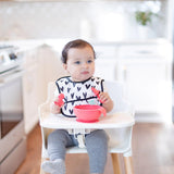 Baby in a high chair wears a heart-print bib and explores Bumkins Red Silicone Chewtensils® with sensory bumps in a bright kitchen.