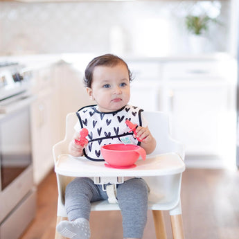 Baby in a high chair wears a heart-print bib and explores Bumkins Red Silicone Chewtensils® with sensory bumps in a bright kitchen.