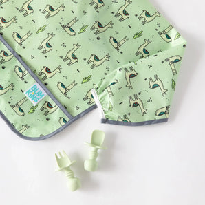 A Bumkins SuperBib® and Sleeved Bib 3-Piece Set: No Prob-Llama, Llamas, Cacti lays flat on a white background near green toddler utensils.