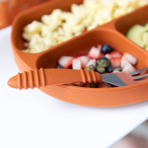 Close-up of an orange divided plate with fruit, pasta, avocado, with a Bumkins Spoon + Fork: Clay resting on the edge.