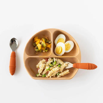 A toddler plate has sliced fruit, halved eggs, and veggie pasta, with Bumkins Spoon + Fork: Clay on a white background.