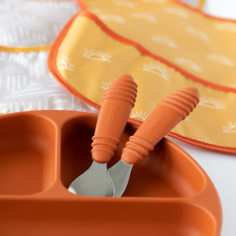 Close-up of Bumkins Spoon + Fork: Clay  in a matching divided plate, with two bibs in the background.
