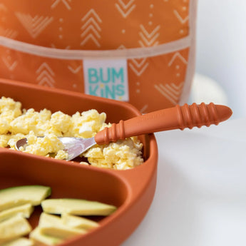 Close-up of a divided orange plate with eggs, avocado, and a Bumkins Spoon + Fork: Clay, with a blurred orange bib in the background.