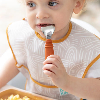 A toddler in a bib holds a Bumkins Spoon + Fork: Clay in their mouth, eating eggs from a brown plate.